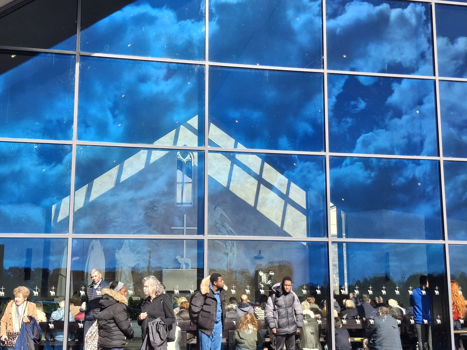 The gable of the Apparition Chapel mirrored in the glass, with the clouds and the pilgrims reflected together