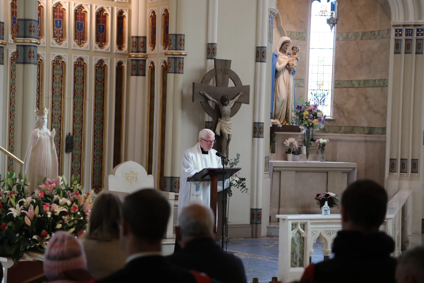 A priest at the lectern, with Our Lady&rsquo;s statue among the flowers beside the sanctuary