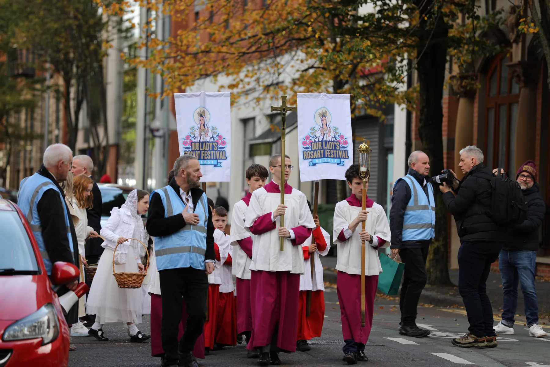 Altar servers carrying the Rosary Festival banners and the processional cross down a Belfast street, stewards alongside