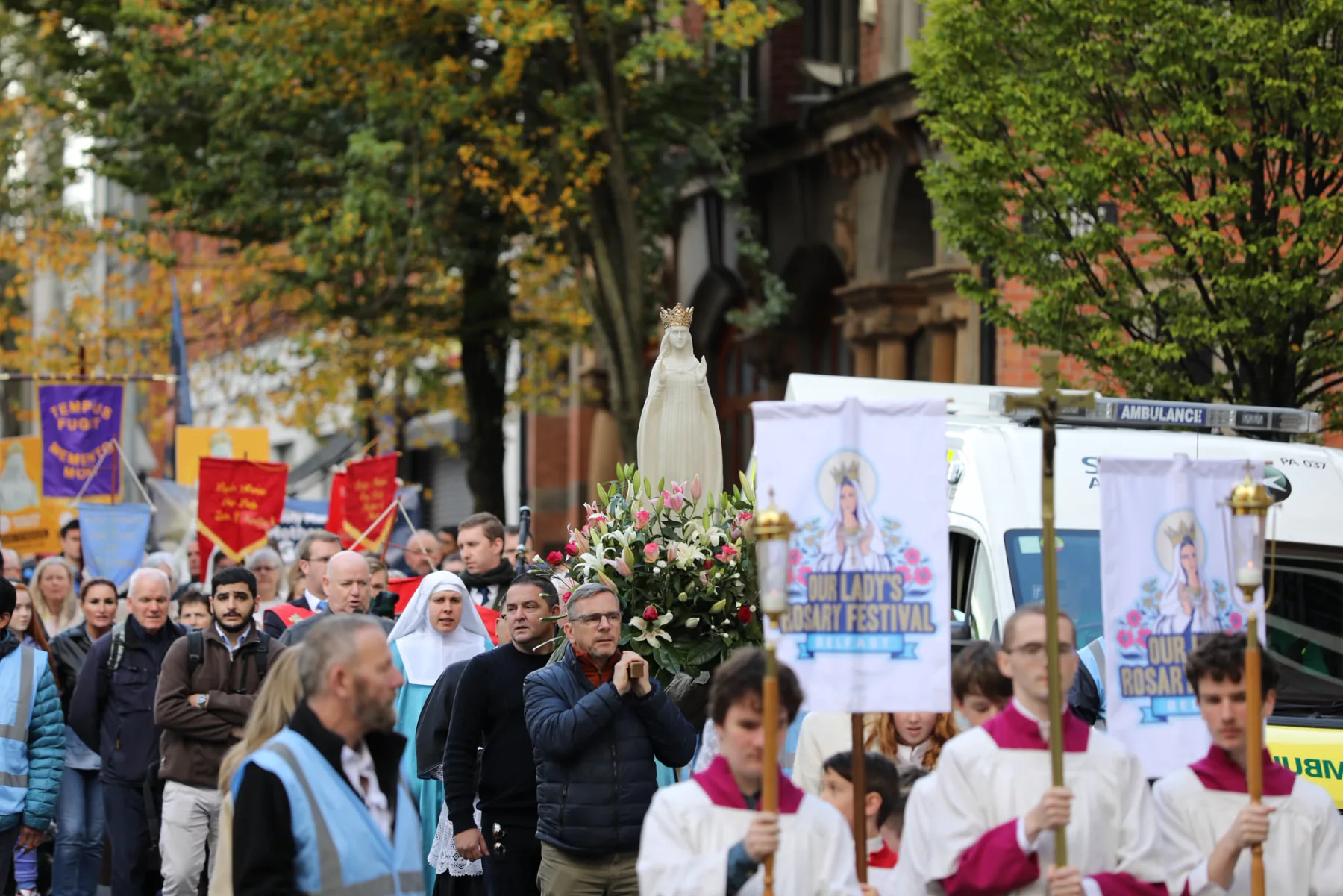 Our Lady&rsquo;s statue carried on a bed of flowers through the crowd, behind the altar servers and banners