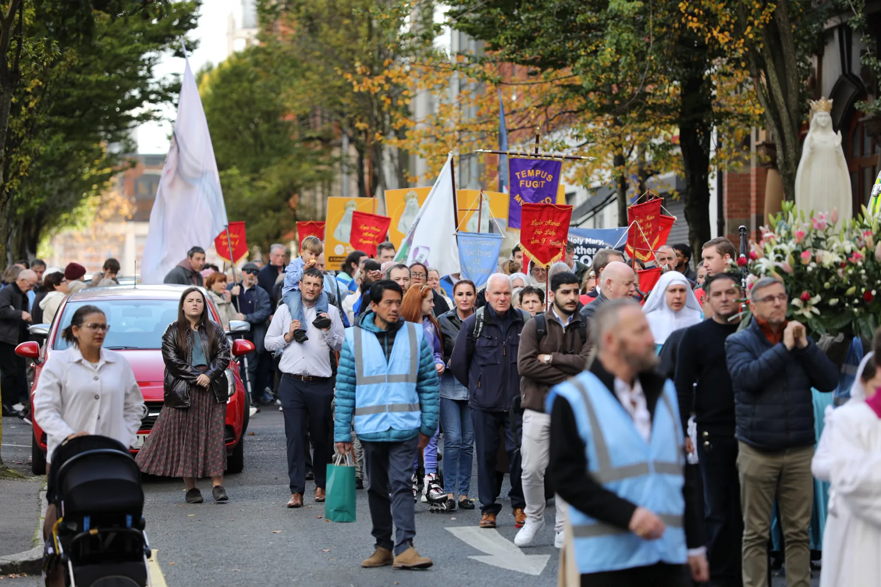 Families, sisters in white habits and stewards walking with the procession, banners raised behind them