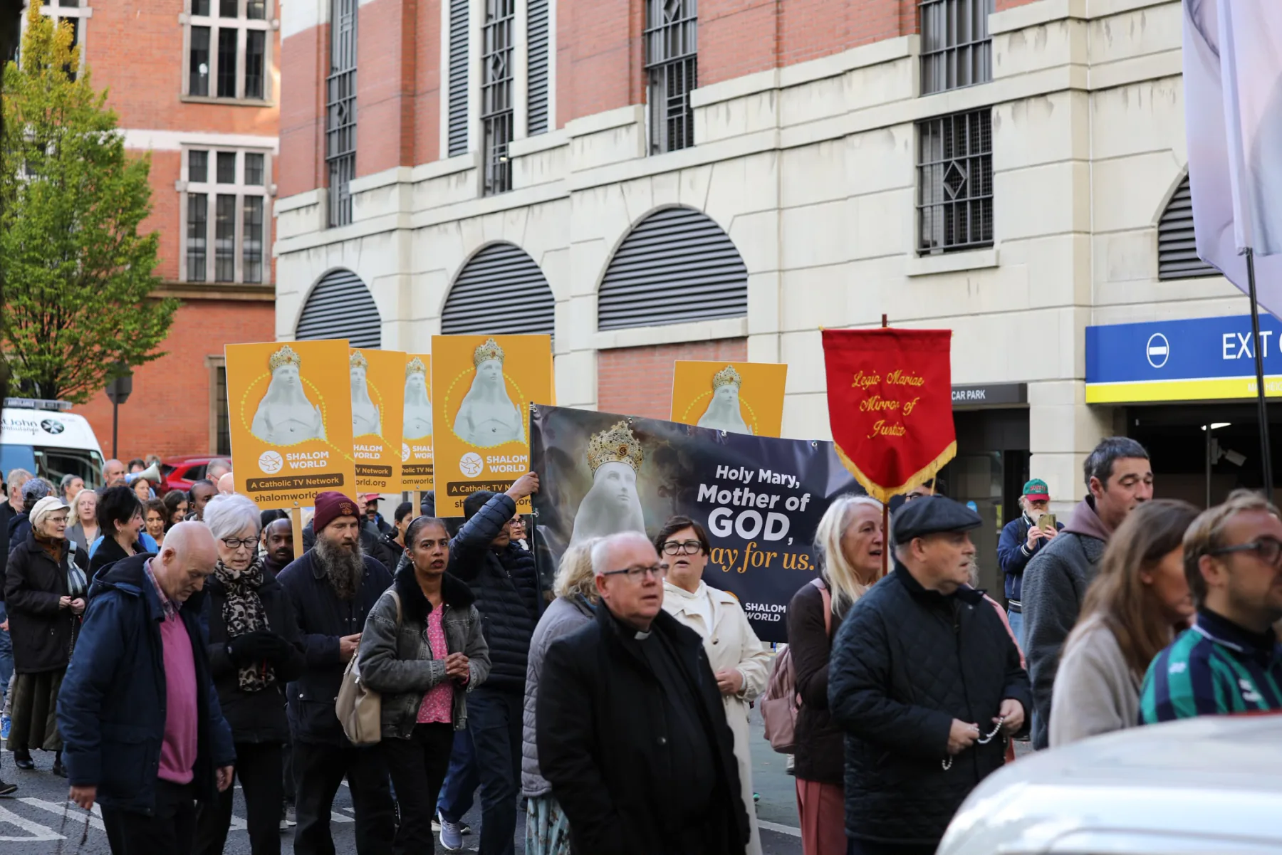 The crowd walking beneath a &lsquo;Holy Mary, Mother of God, pray for us&rsquo; banner and the red parish banners
