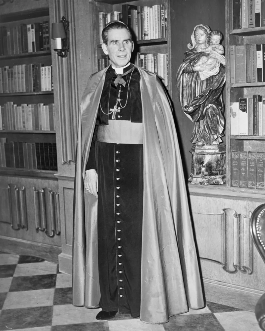 Archbishop Fulton J. Sheen in cape and pectoral cross, standing in a library beside a carved statue of Our Lady holding the Child Jesus