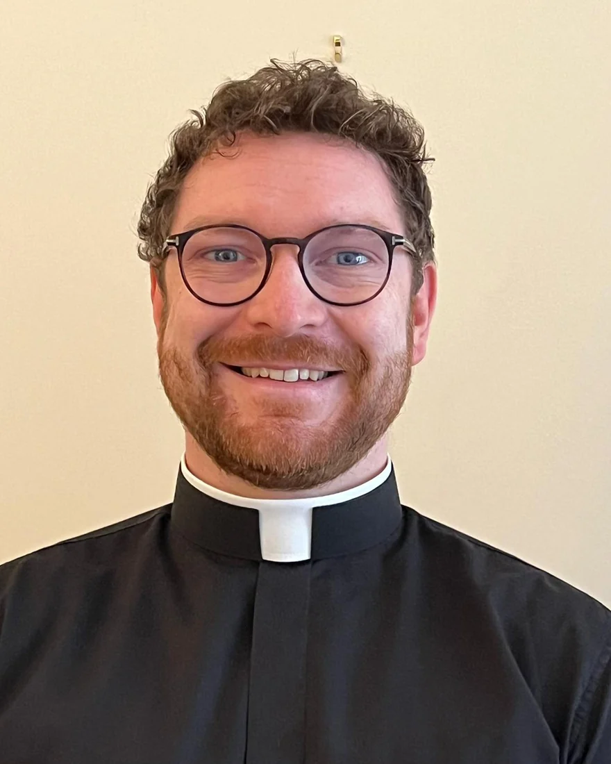 Fr John O&rsquo;Laverty, wearing a clerical collar and glasses, smiling at the camera